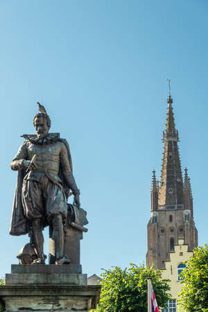 Bruges, Flanders, Belgium -  June 17, 2019: Simon Stevin statue with spire of OLV Cathedral in back under blue sky. Some green foliage. Pigeon on his head.のeditorial素材