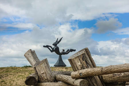 Knokke-Heist, Flanders, Belgium -  June 16, 2019: Knokke-Zoute part of town. Jumping hare statue on brown-green dune behind pile of wood. Captured in  blue and white cloudscape.のeditorial素材