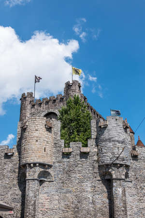 Gent, Flanders, Belgium -  June 21, 2019: Gray stone tower and ramparts of Gravensteen, historic medieval castle of city against blue sky with white clouds. Flags on top, green foliage.のeditorial素材