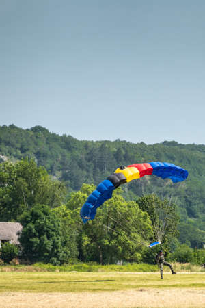 Han-sur-Lesse, Belgium - June 25, 2019: Belgian soldier on parachute with Belgian flag colors lands on field with forested hills and blue sky in back.のeditorial素材