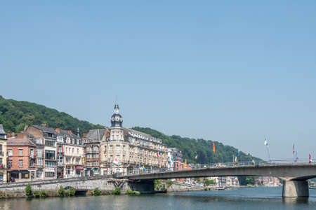 Dinant, Belgium - June 26, 2019: Charles de Gaule bridge spans Meuse River under blue sky. Left bank buildings with historic towered corner building. flags and green foliage.のeditorial素材