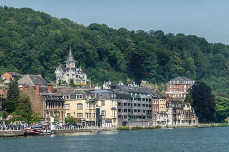 Dinant, Belgium - June 26, 2019: Left bank, north flow of Meuse River with buildings backed by green foliage and blue sky. Tower on mansion in forest foliage.のeditorial素材