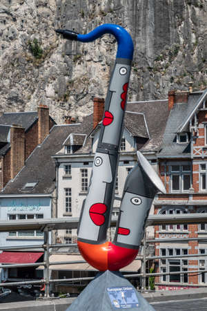Dinant, Belgium - June 26, 2019: Red, gray and blue saxophone statue to honor the European Union on Charles de Gaulle bridge. Business and houses in back against gray rock cliff.のeditorial素材