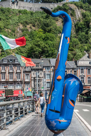 Dinant, Belgium - June 26, 2019: blue saxophone statue on Charles de Gaulle bridge. Business and houses in back. Country flags and people.のeditorial素材