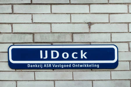 Amsterdam, the Netherlands - June 30, 2019: White on deep blue street sign naming IJDOCK and sponsored by real estate company, placed on white brick wall.のeditorial素材