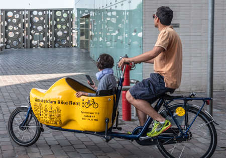 Amsterdam, the Netherlands - June 30, 2019: Closeup of bakfiets, front-trunk bike to transport a load, a child here. Man pedaling as seen on IJdok. Trunk in shape of yellow clog.のeditorial素材