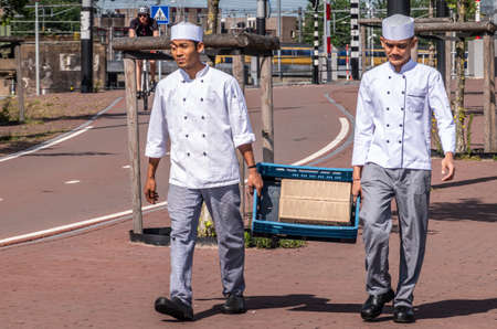 Amsterdam, the Netherlands - June 30, 2019: Two young male kitchen helpers in uniform carry blue crate with Lamb meat box along street near IJdok.のeditorial素材