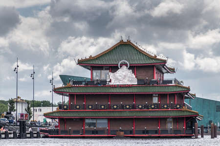 Amsterdam, the Netherlands - July 1, 2019: Red-green-brown short facade with huge white Buddha statue of Sea Palace floating restaurant at Oosterdokskade under heavy cloudscape.のeditorial素材