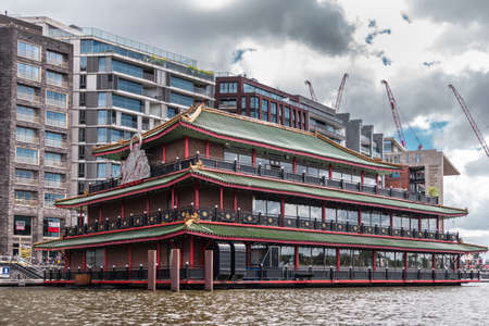 Amsterdam, the Netherlands - July 1, 2019: Red-green-brown long facade with huge white Buddha statue of Sea Palace floating restaurant at Oosterdokskade under heavy cloudscape.のeditorial素材