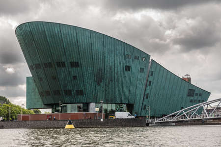 Amsterdam, the Netherlands - July 1, 2019: Modern boat hull building housing the NEMO science museum on border of Oosterdok in black water under havey rainy cloudscape.のeditorial素材