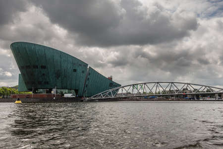 Amsterdam, the Netherlands - July 1, 2019: Modern boat hull building housing the NEMO science museum on border of Oosterdok in black water under havey rainy cloudscape.のeditorial素材