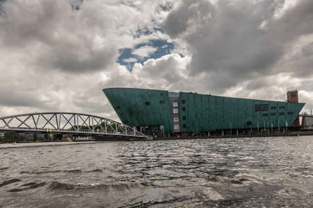 Amsterdam, the Netherlands - July 1, 2019: Modern boat hull building housing the NEMO science museum on border of Oosterdok in black water under havey rainy cloudscape.のeditorial素材
