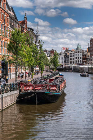 Amsterdam, the Netherlands - July 1, 2019: Dark flat barge is used as floating bike parking on Singel canal under blue-gray cloudscape. Munt tower spire. Glass houses of floating flower market on other side.のeditorial素材