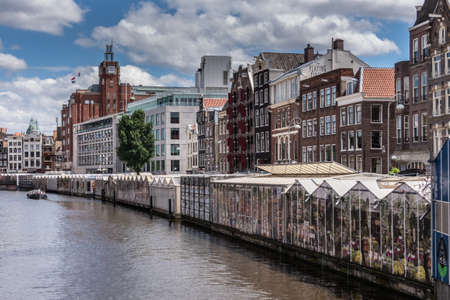 Amsterdam, the Netherlands - July 1, 2019: Long row of glasshouses built on barges moored on Singel Canal and housing flower market. Back is row of houses under blue cloudscape,のeditorial素材