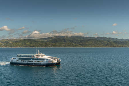 Manoc-Manoc, Boracay, Philippines - March 4, 2019: White and blue Fast Cat motorized modern long distance ferry sails on sea under blue sky. Mountains with windmills on horizon.のeditorial素材