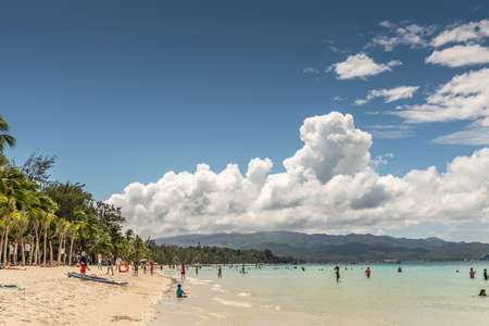 Balabag, Boracay Island, Philippines - March 4, 2019: White beach shows sand and azure sea with wall of palm trees. People in water. Cloudscape over Malay Island.のeditorial素材