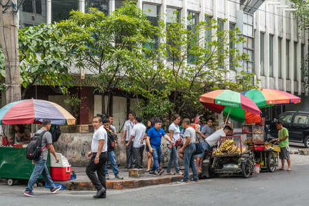 Manila, Philippines - March 5, 2019: Three ambulant food vendors with push cart or tricycle and umbrellas on corner of street under office building. Customers in line.のeditorial素材