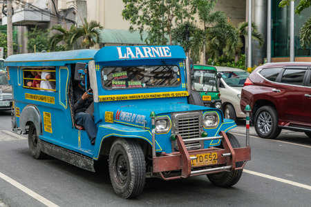Manila, Philippines - March 5, 2019: Sky blue long Isuzu Jeep Public Transport called Laarnie in street. Side and front view. People inside. Green foliage. Other car and gray buildings.のeditorial素材