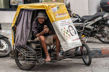Manila, Philippines - March 5, 2019: Smoking older tricycle cab driver waits inside his vehicle for customers along street.のeditorial素材