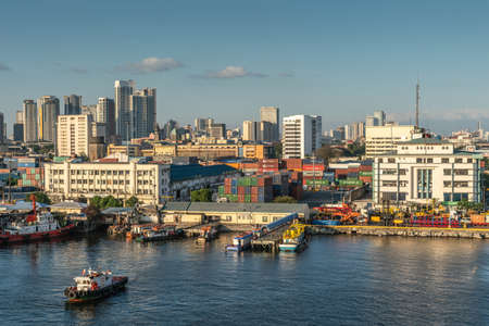 Manila, Philippines - March 5, 2019: South Harbor evening twilight. Harbor management buildings on shore behind cranes, trucks, containers and boats on the water. Tall buildings on horizon.のeditorial素材