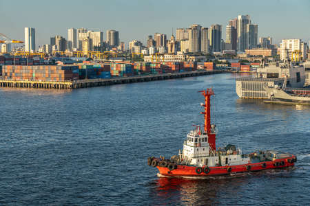 Manila, Philippines - March 5, 2019: South Harbor evening twilight. Red tug boat up front. Navy vessels, a pier full of shipping containers in back. Line of tall buildings on horizon. light blue sky.のeditorial素材