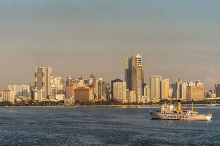 Manila, Philippines - March 5, 2019: South Harbor evening twilight shines on tall buildings such as Eton and Birch tower with Amosup rescue ships on dark blue sea water.のeditorial素材