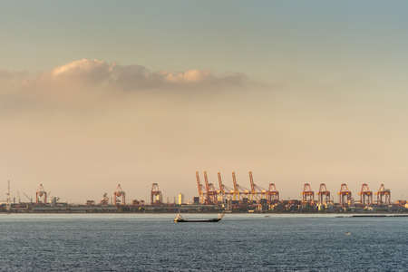 Manila, Philippines - March 5, 2019: Evening twilight shines on multiple red container cranes in container terminal. Ship passes by on darker blue water.のeditorial素材