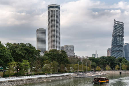 Singapore - March 20, 2019: Sightseeing boat on Singapore River with Swissotel the Stamford and JW Marriott hotel towers in back. Esplanade Park adds green foliage under cloudscape.のeditorial素材