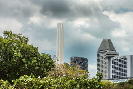Singapore - March 20, 2019: Beige tall war memorial obelisk towering over green foliage of War Memorial Park under cloudscape. To the side, skyscrapers such as Pan Pacific.のeditorial素材
