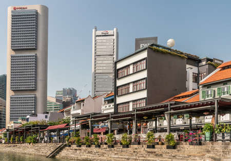 Singapore - March 21, 2019: Along Singapore River. A few financial district towers behind smaller houses now converted into restaurants and bars under blue sky. Plants add green.のeditorial素材