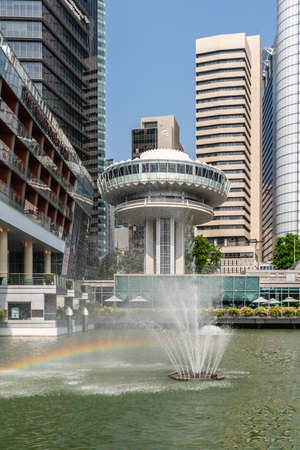 Singapore - March 21, 2019: On Marina waters. Clifford Square with rainbow through fountain and towers of Financial District in back. Customs House tower. Some blue sky and green foliage.のeditorial素材