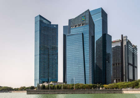 Singapore - March 21, 2019: On Marina waters. Gorup of blue skyscrapers of financial district nearest to the bay under light blue sky. Green foliage and water in front.のeditorial素材