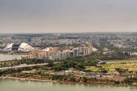 Singapore - March 21, 2019: Shot from Sands roof. Birds eye view on open National Stadium with housing and hundreds of skyscrapers in wide area. Silver sky and greenish bay water.のeditorial素材