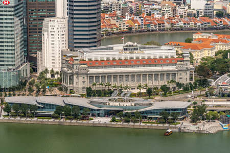 Singapore - March 21, 2019: Shot from Sands roof. Birds eye closeup of historic The Fullerton Hotel with its modern bay section up front on greenish water. River and shore buildings in back.のeditorial素材