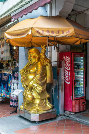 Singapore - March 22, 2019: Chinatown. Golden laughting Buddha statue used by Venhonia Charity to collect funds in front of shop in Sago Street. Red Coca-cola vending machine in back.のeditorial素材