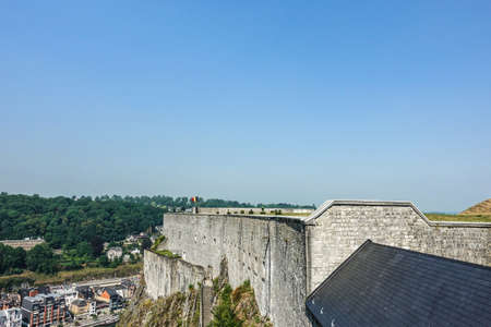 Dinant, Belgium - June 26, 2019: Gray stone south ramparts of Citadelle fortification with Belgian Flag at top under blue sky. Horizon is forest and down the Meuse River and cityscape.のeditorial素材