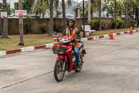 Si Racha, Thailand - March 16, 2019: Man with orange vest rides red Honda motorcycle on Ko Loi Island. Gray street surface and green foliage in back.のeditorial素材
