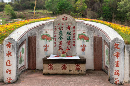 Bang Phra, Thailand - March 16, 2019: Chao Pho Khao Chalak Chinese Cemetery. Closeup of gray tombstone with painted Mandarin letters, red roses, incense sticks in a cup, and yellow and red flowers on hill.のeditorial素材