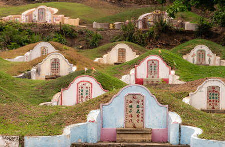 Bang Phra, Thailand - March 16, 2019: Chao Pho Khao Chalak Chinese Cemetery. Group of family graves with light blue one in focus. Mandarin lettering, green hills and foliage.のeditorial素材