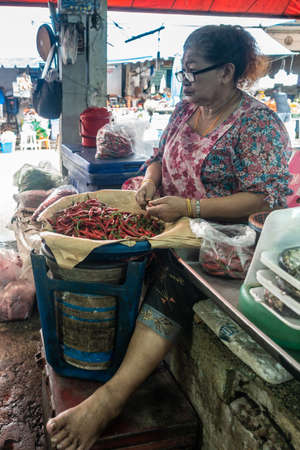 Chon Buri, Thailand - March 16, 2019: Nong Mon market. Sitting woman with big glasses cleans pile of red chilli peppers in her fish selling booth.のeditorial素材