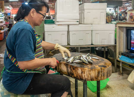 Chon Buri, Thailand - March 16, 2019: Nong Mon market. Female vendor cleans small gray fishes on filthy wooden table while watching TV in her booth.のeditorial素材