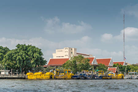 Bangkok city, Thailand - March 17, 2019: Chao Phraya River. Group of yellow and blue river maintenance boats with cranes and dredging equipement in front of red roofed Departement of Law buildings.のeditorial素材