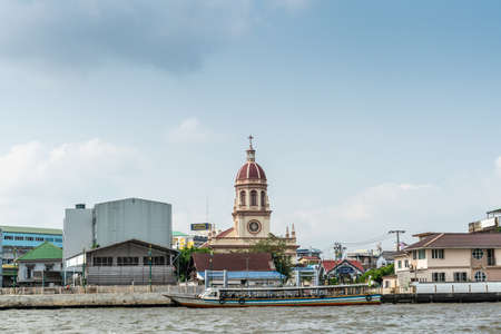 Bangkok city, Thailand - March 17, 2019: Chao Phraya River. Red domed beige tower of Santa Cruz Catholic church with cross on top behind boats on river and other buildings under light blue sky.のeditorial素材