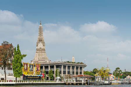 Bangkok city, Thailand - March 17, 2019: Chao Phraya River. Royal Thai Navy Headquarter buildings with colorful posters of Royalty with prangs of Temple of Dawn in back under light blue cloudscape.のeditorial素材