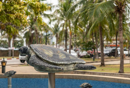 Si Racha, Thailand - March 16, 2019: Bronze statue of turtle on pole above blue water pond on Ko Loi Island. Green foliage in back, and cars.のeditorial素材