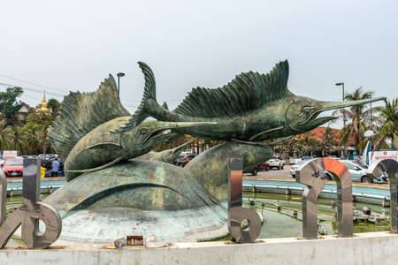Si Racha, Thailand - March 16, 2019: Koh Loy Park fountain with large bronze statues of swordfishes under blue sky with green foliage and red roof in back. Cars and people.のeditorial素材