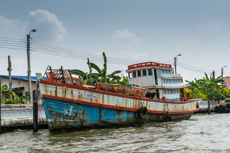 Bangkok city, Thailand - March 17, 2019: Bangkok Noi Canal. Closeup of blue-red-white decent tranport vessel with large cabin and ribble on deck docked at quay under blue sky, Green foliage adds color.のeditorial素材