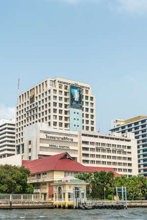 Bangkok city, Thailand - March 17, 2019: Chao Phraya River. White with black text and blueish poster on top of Siriraj Hospital building under blue sky. Green foliage and red roof of smaller building up front.のeditorial素材