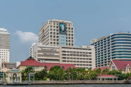 Bangkok city, Thailand - March 17, 2019: Chao Phraya River. White with black text and blueish poster on top of Siriraj Hospital building under blue sky. Green foliage and red roofs of smaller buildings up front.のeditorial素材