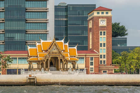 Bangkok city, Thailand - March 17, 2019: Chao Phraya River. Red brick clock tower of old railway station now Siriraj Bimuksthan Museum with Buddhist shrine up front. Modern tall buildings in back and some green foliage.のeditorial素材